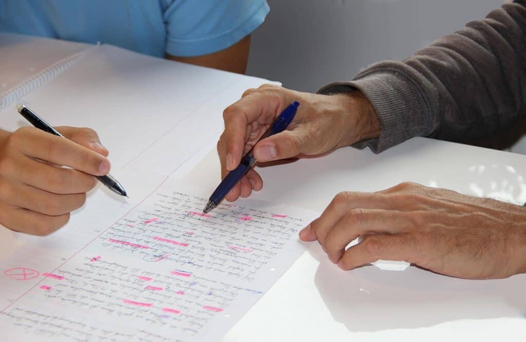 Zwei Personen begutachten ein handgeschriebenes Dokument mit rosafarbenen Unterstreichungen und Anmerkungen; die eine zeigt mit einem Stift, während die andere schreibt, wobei die Notizbücher auf dem Tisch sichtbar sind.