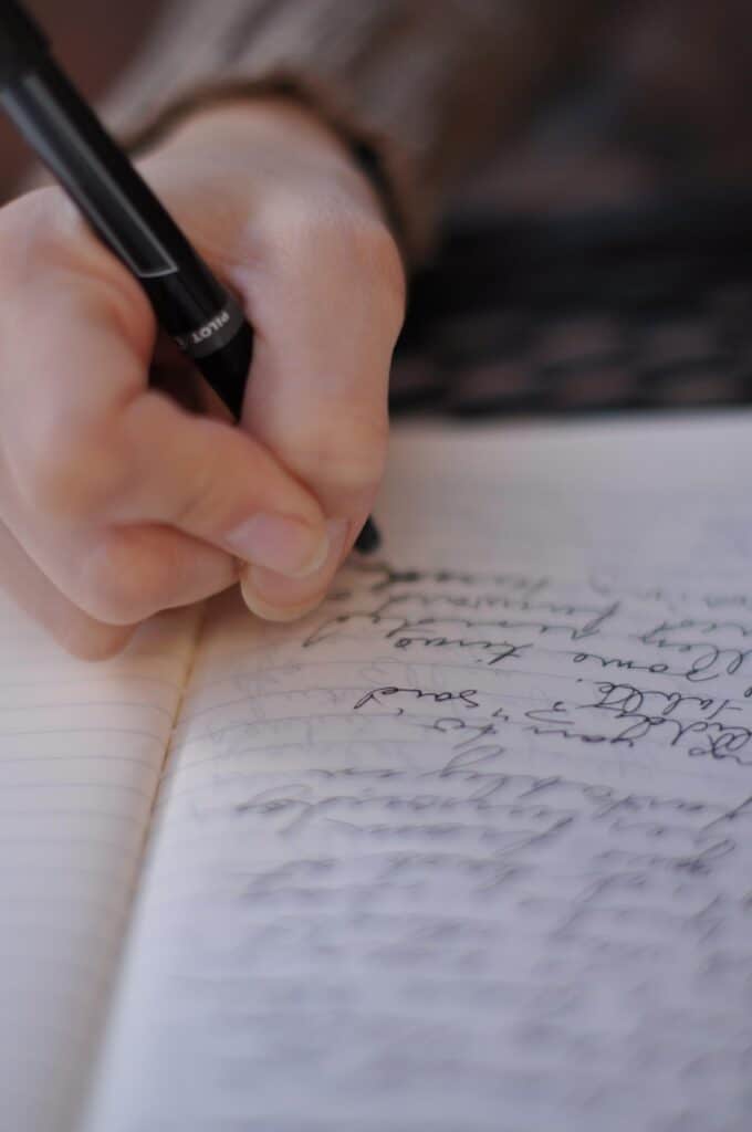 Eine Person, die mit einem schwarzen Stift in ein liniertes Notizbuch schreibt, wobei der handschriftliche Text auf der Seite sichtbar ist.