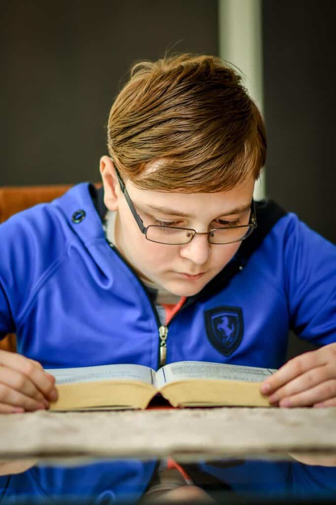 Ein Junge mit Brille und blauer Jacke liest aufmerksam an einem Tisch ein Buch.